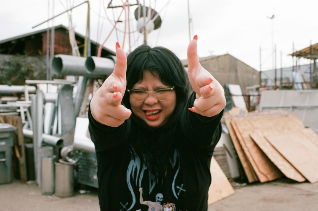 a woman wearing glasses with black hair and bangs strikes a pose with her hands shaped as finger guns toward the camera. in the background is construction materials like wooden planks and barrels found at a recycling depot.